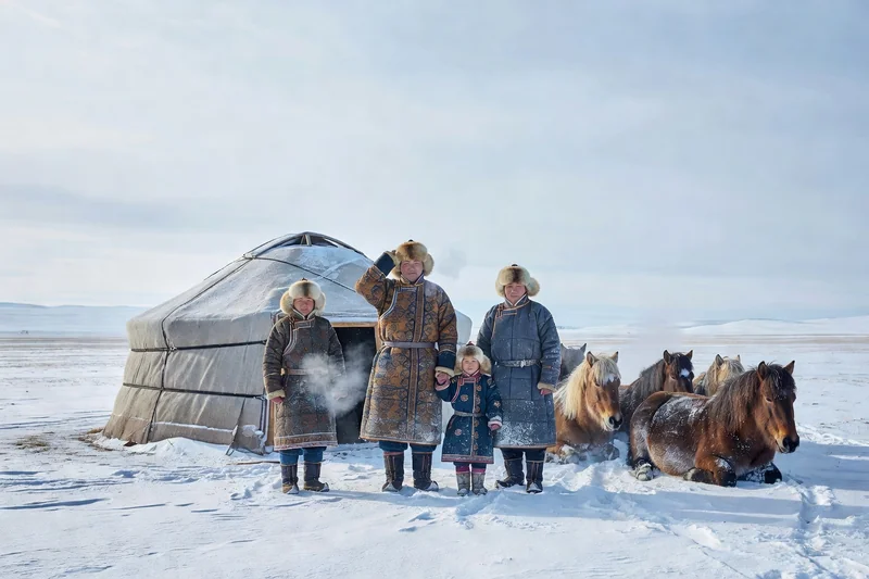 Famille nomade mongole en vêtements d'hiver traditionnels devant sa yourte, avec des chevaux au repos dans la steppe enneigée